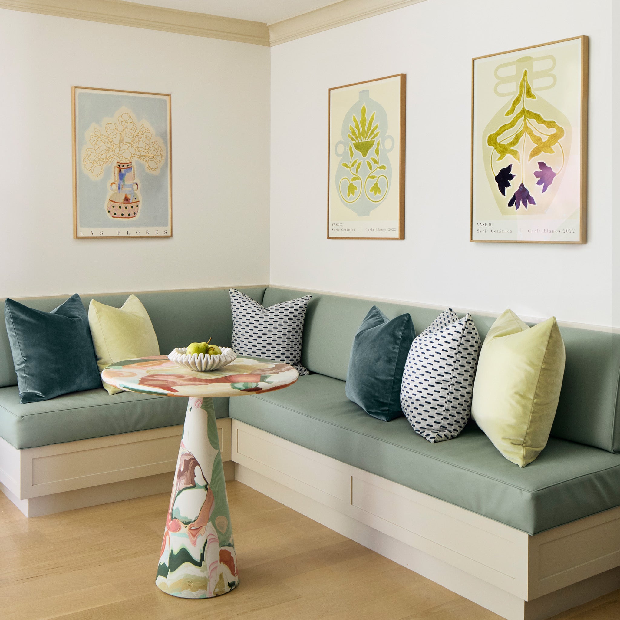 sage green kitchen bench with chenille and woven jacquard mint green geometric, pink, and teal pillows on it with floral artwork hung on the walls above it and a multicolored table 