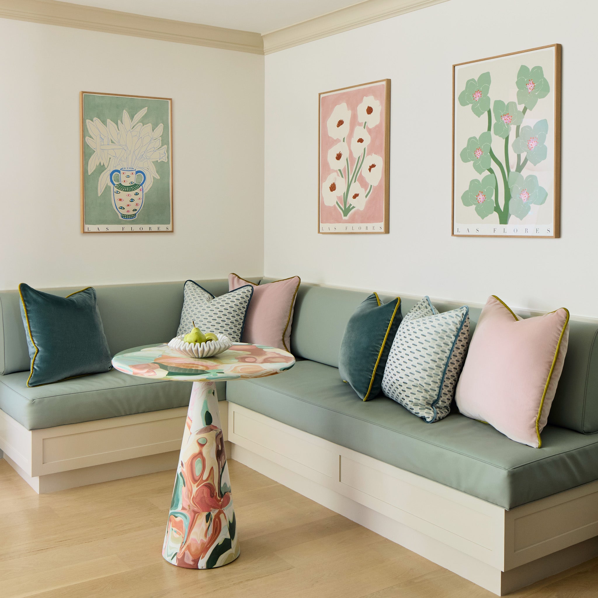 sage green kitchen bench with chenille and woven jacquard mint green geometric, pink, and teal pillows on it with floral artwork hung on the walls above it and a multicolored table 