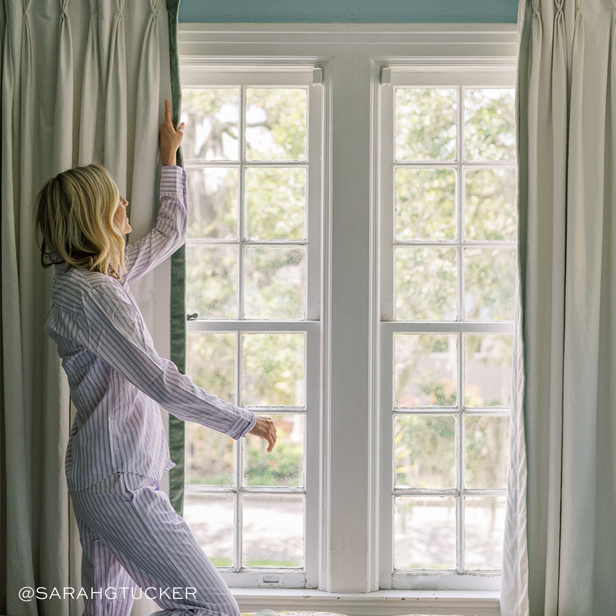 Woman adjusting Flour made-to-order curtains in soft neutral tones, manufactured in the USA with quick turnaround and quality design