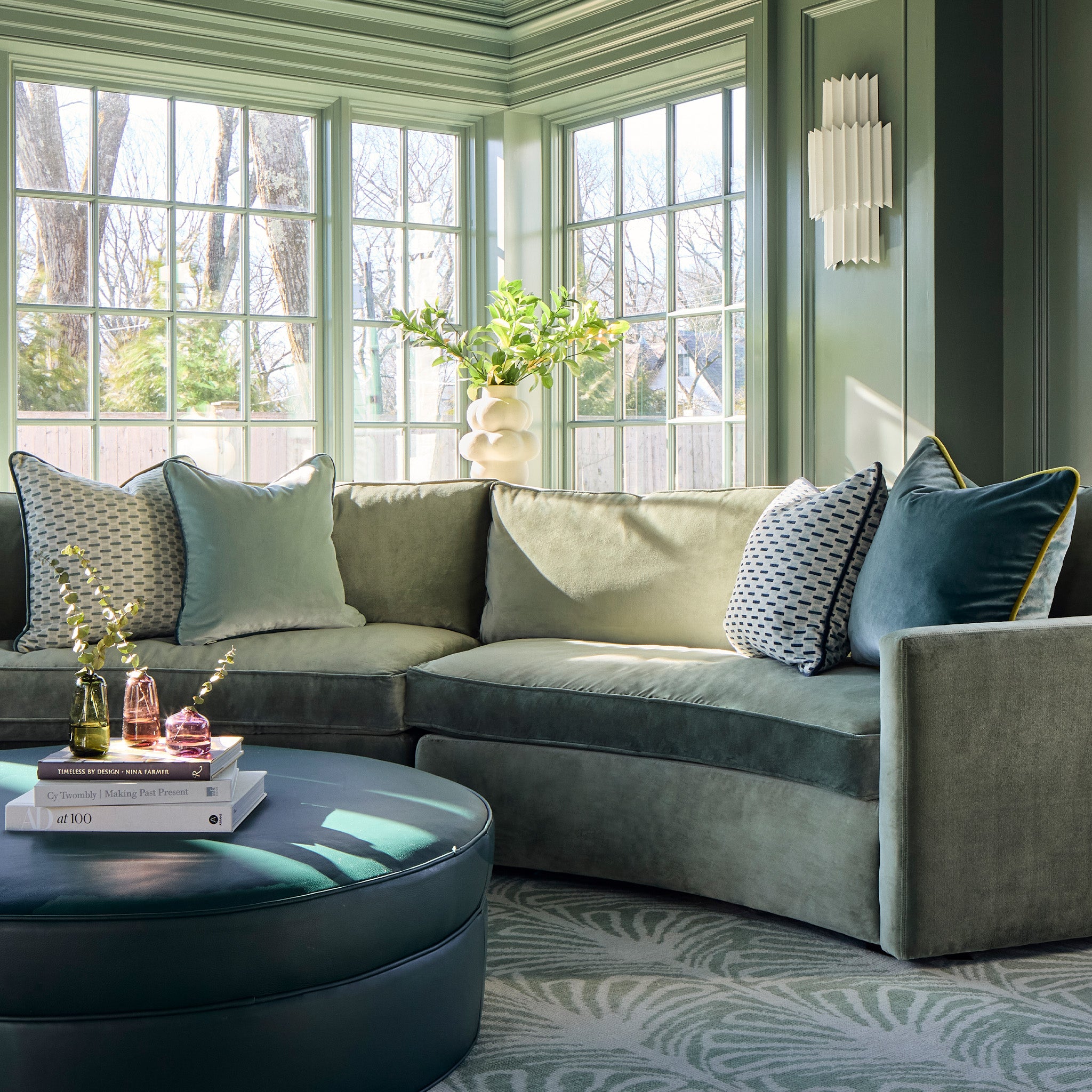 green couch with chenille and woven jacquard mint green geometric, sage green, and teal blue pillows on it with a teal blue ottoman stacked with books in a room painted green