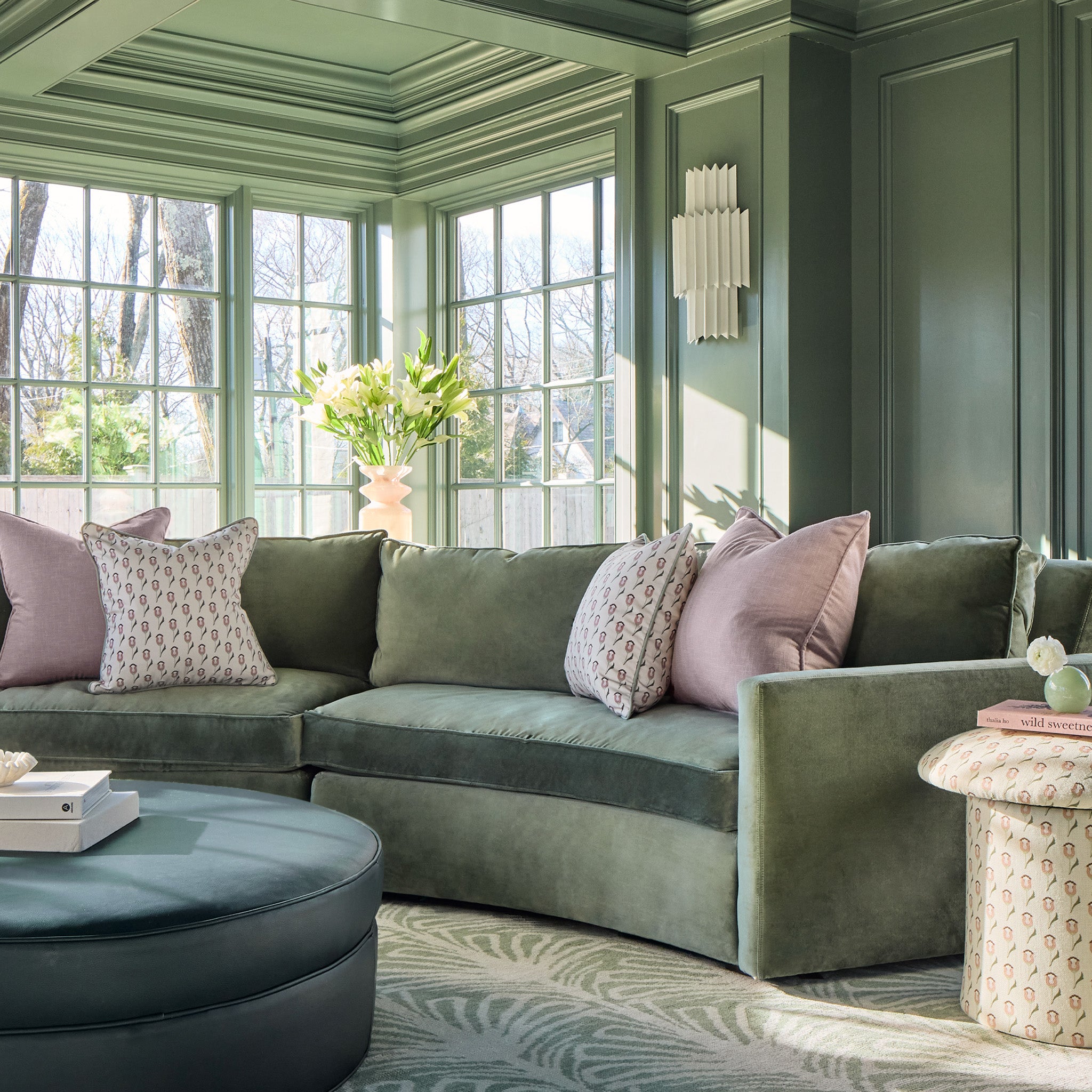abstract floral pink and green pillows on a green couch with a blue ottoman with books stacked on it and green painted walls