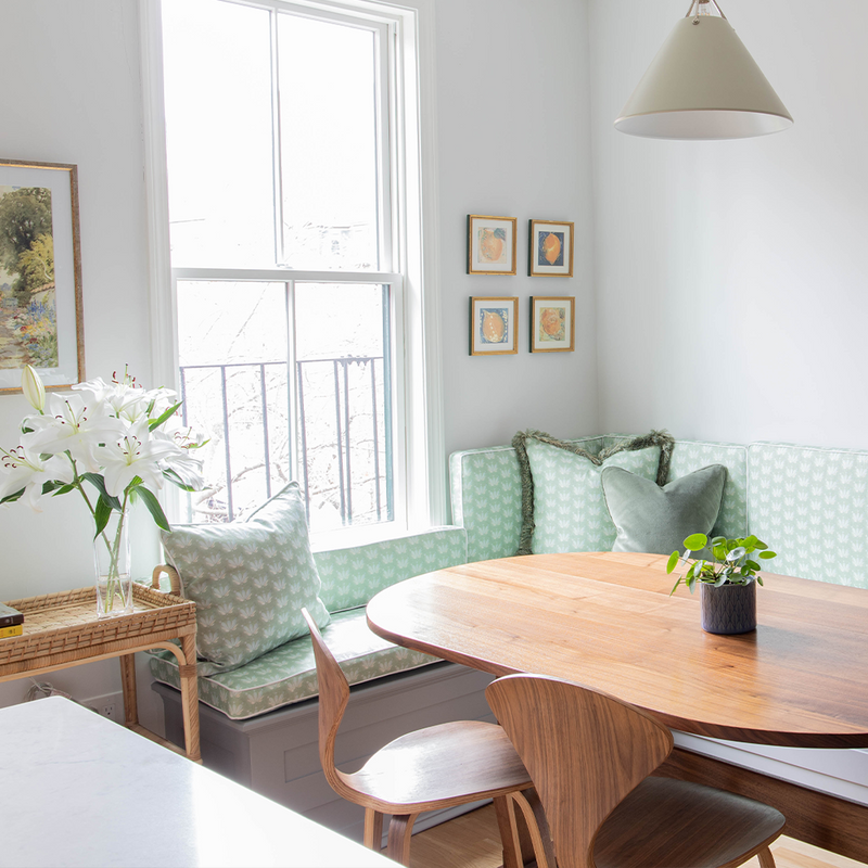 Blue & Green Floral Drop Repeat pattern cushion on a kitchen bench with Blue & Green Floral Drop Repeat pillows on it with an illuminated window behind the bench and a wooden table in front of the bench 